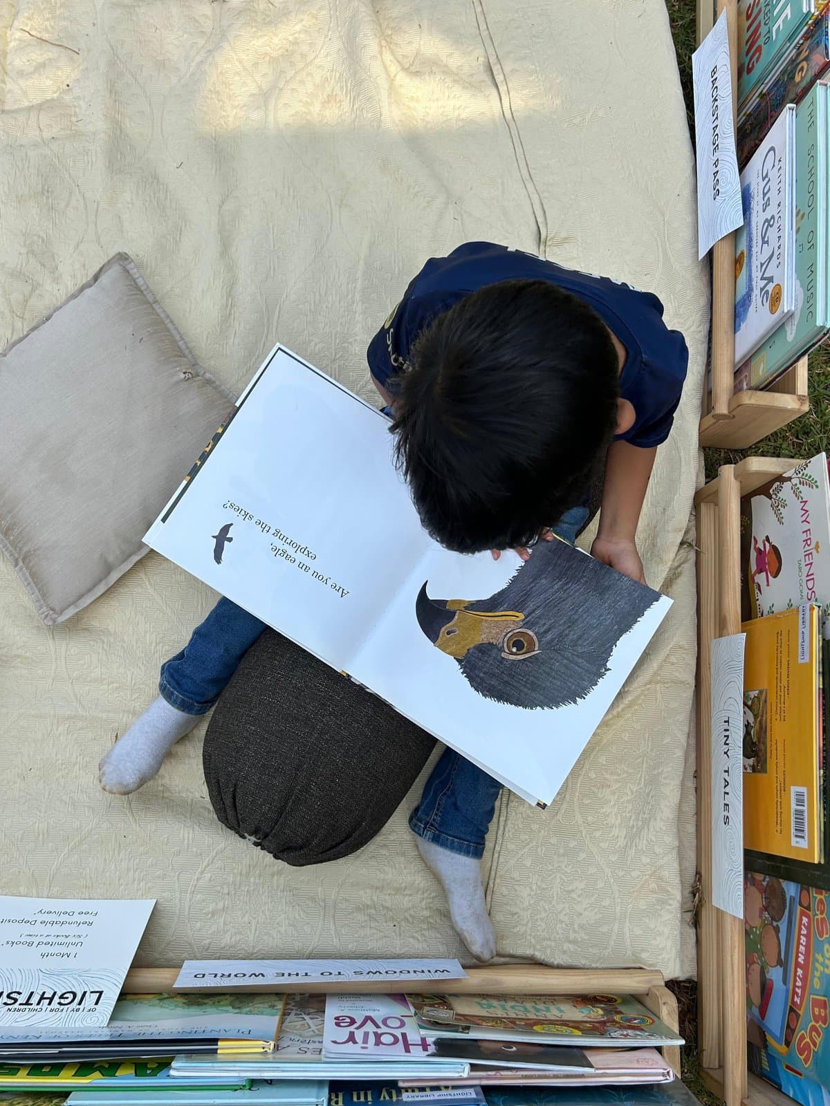 A child sitting on a picnic blanket at a Lightship pop-up, absorbed in an open picture book, surrounded by shelves of curated books